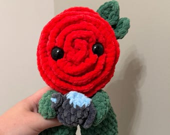 May include: A plush toy shaped like a red rose flower with a green stem and leaves. The rose head features two black plastic eyes. The toy is holding a small, dark gray, textured object with a light blue top between its green crocheted arms.