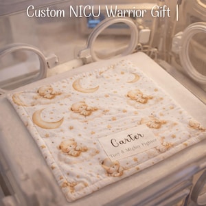 May include: A white baby blanket with a celestial design featuring teddy bears, moons, and stars. The blanket has a personalized label that reads "Carter Tiny & Mighty Fighter". The blanket is inside a NICU incubator.