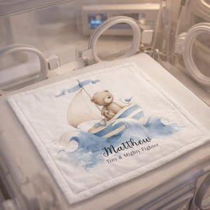 May include: A white baby blanket with a watercolor illustration of a teddy bear sailing in a boat on blue waves. The blanket has the name "Matthew" and the text "Tiny & Mighty Fighter" printed on it.