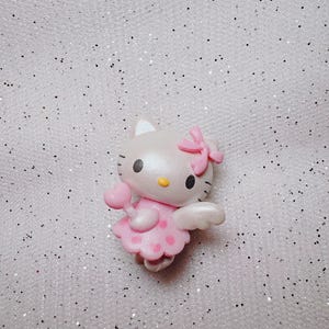 May include: A pink and white Hello Kitty figurine with wings and a heart-shaped wand. She is wearing a pink dress with white polka dots and a pink bow.