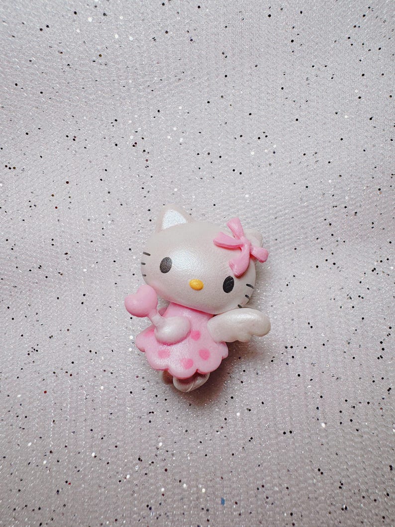 May include: A pink and white Hello Kitty figurine with wings and a heart-shaped wand. She is wearing a pink dress with white polka dots and a pink bow.