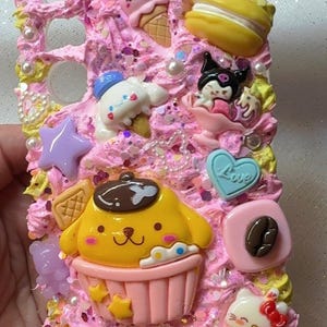 May include: A pink and yellow phone case decorated with various kawaii characters and objects, including a cupcake, ice cream cone, a bow, a star, a cloud, a coffee bean, and a Hello Kitty character. The case is covered in pink and white frosting with sprinkles and pearls.