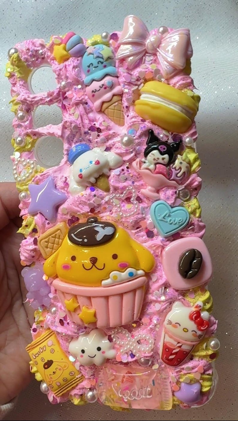 May include: A pink and yellow phone case decorated with various kawaii characters and objects, including a cupcake, ice cream cone, a bow, a star, a cloud, a coffee bean, and a Hello Kitty character. The case is covered in pink and white frosting with sprinkles and pearls.
