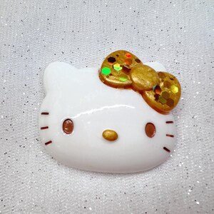 Peut inclure: Figurines en c&eacute;ramique blanche d'un chat de dessin anim&eacute; avec un n&oelig;ud en paillettes dor&eacute;es. Le chat a des yeux bruns et un nez rose.