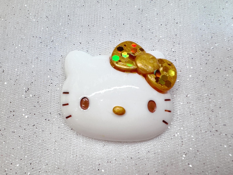 Peut inclure: Figurines en c&eacute;ramique blanche d'un chat de dessin anim&eacute; avec un n&oelig;ud en paillettes dor&eacute;es. Le chat a des yeux bruns et un nez rose.