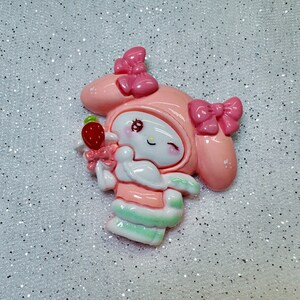 Peut inclure: Personnage de dessin anim&eacute; rose et blanc avec un n&oelig;ud rose et une fraise dans la main. Le personnage porte une tenue rose et blanche et a un &oelig;il qui cligne.