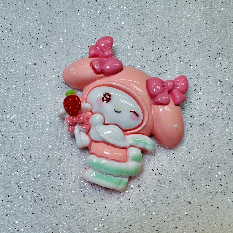 Peut inclure: Personnage de dessin anim&eacute; rose et blanc avec un n&oelig;ud rose et une fraise dans la main. Le personnage porte une tenue rose et blanche et a un &oelig;il qui cligne.
