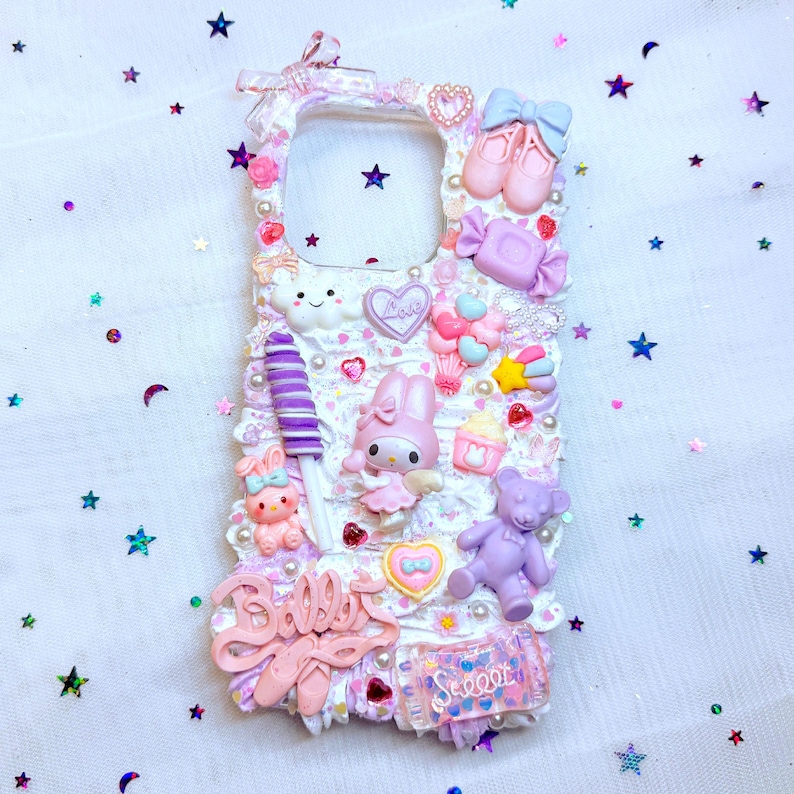 May include: A white phone case decorated with pastel pink, purple, and blue 3D embellishments. The case features a cloud, a heart, a lollipop, a bow, a candy cane, a star, and a ballerina shoe. The text "Sweet" and "Baller" is visible on the case.