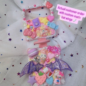 May include: A custom-made phone case with a variety of colorful, decorative elements. The case features a pink and white base, adorned with candies, hearts, stars, and a pair of purple glitter bat wings. The word "Sweet" is written on the case.