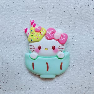 May include: A white Hello Kitty character with pink bow and yellow and pink sprinkles on her head, sitting in a light blue cup with gold accents.