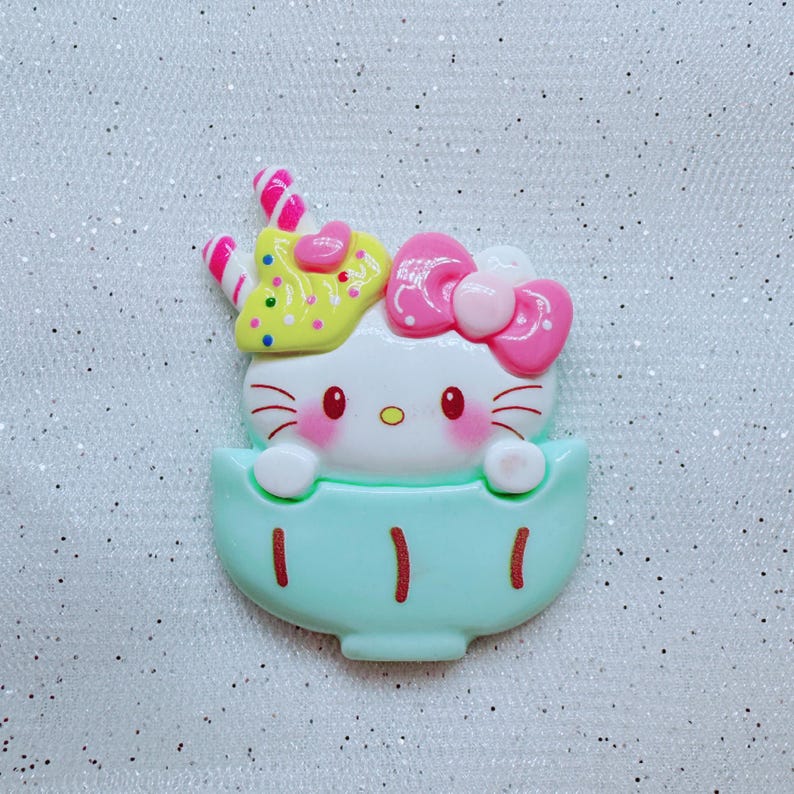 May include: A white Hello Kitty character with pink bow and yellow and pink sprinkles on her head, sitting in a light blue cup with gold accents.