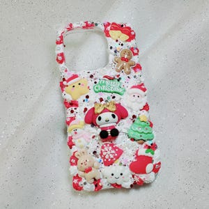 K&ouml;nnte beinhalten: Eine wei&szlig;e Handyh&uuml;lle, die mit weihnachtlichen Figuren und Glitzer verziert ist. Die H&uuml;lle zeigt einen Lebkuchenmann, einen Schneemann, einen Weihnachtsbaum, einen Strumpf und eine Wolke mit einem Smiley. Der Text "Merry Christmas" ist auf der H&uuml;lle aufgedruckt.
