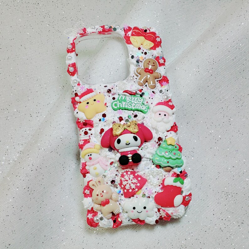 K&ouml;nnte beinhalten: Eine wei&szlig;e Handyh&uuml;lle, die mit weihnachtlichen Figuren und Glitzer verziert ist. Die H&uuml;lle zeigt einen Lebkuchenmann, einen Schneemann, einen Weihnachtsbaum, einen Strumpf und eine Wolke mit einem Smiley. Der Text "Merry Christmas" ist auf der H&uuml;lle aufgedruckt.