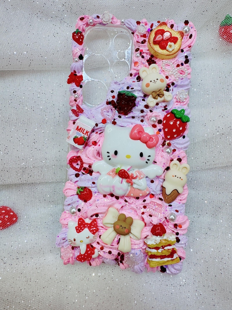Peut inclure: Une coque de t&eacute;l&eacute;phone rose et blanche d&eacute;cor&eacute;e de Hello Kitty, de fraises et d'autres personnages mignons. La coque est recouverte de gla&ccedil;age rose et blanc et de p&eacute;pites.
