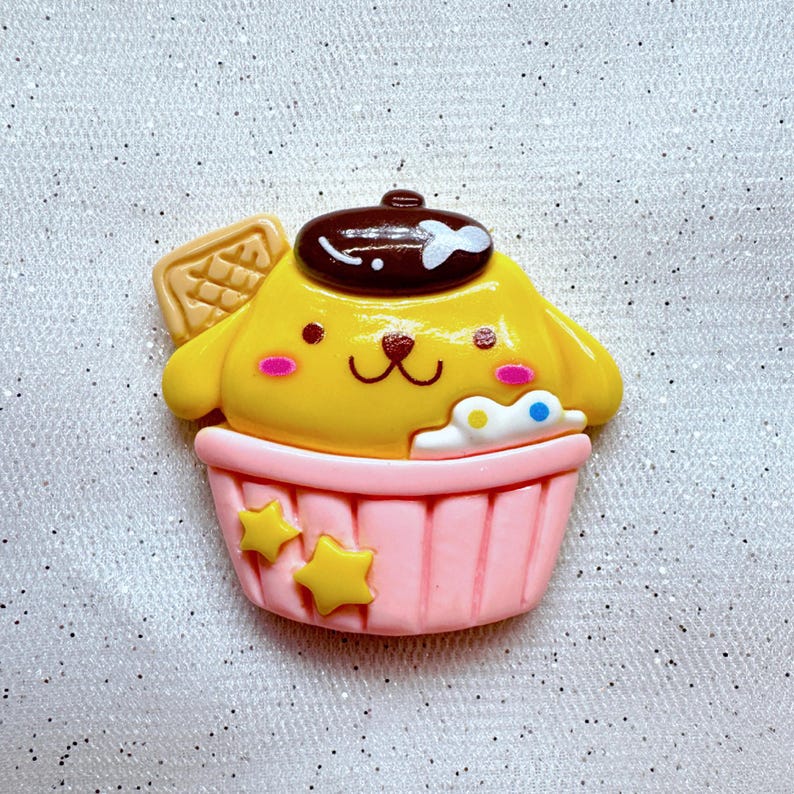 May include: A yellow cupcake with a brown beret and a waffle cone on top. The cupcake is decorated with white frosting and colorful sprinkles. The cupcake is in a pink cupcake liner with yellow stars.