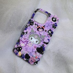 May include: A lilac phone case adorned with charms and decorations. The case showcases a large Kuromi character, alongside other character charms, bows, hearts, and beads. The case is covered in glitter and has a textured finish.