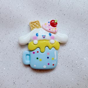 May include: A blue and white ceramic mug with a white dog character with pink cheeks and blue eyes. The dog is wearing a pink and white hat with a red cherry on top. The mug is decorated with yellow icing and colorful sprinkles.