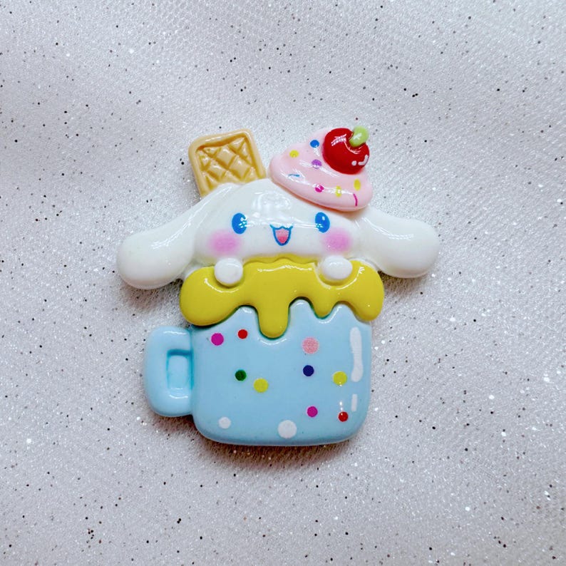 May include: A blue and white ceramic mug with a white dog character with pink cheeks and blue eyes. The dog is wearing a pink and white hat with a red cherry on top. The mug is decorated with yellow icing and colorful sprinkles.