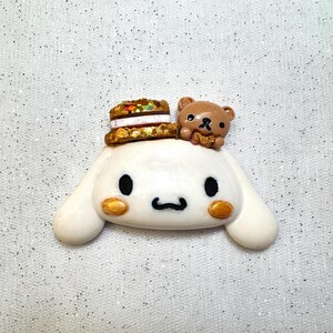 Peut inclure: Une figurine de chien blanc avec des yeux bruns et une figurine d'ours brun sur la t&ecirc;te. Le chien porte un chapeau de g&acirc;teau brun et dor&eacute;.