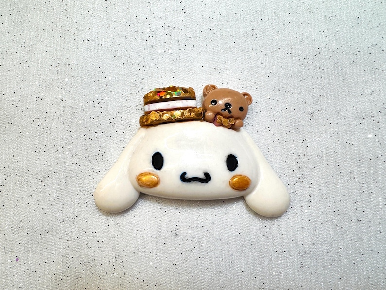 Peut inclure: Une figurine de chien blanc avec des yeux bruns et une figurine d'ours brun sur la t&ecirc;te. Le chien porte un chapeau de g&acirc;teau brun et dor&eacute;.