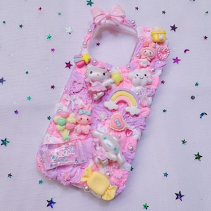 Könnte beinhalten: Ein rosa-lila Handyhülle, verziert mit verschiedenen Kawaii-Charms. Die Hülle zeigt Hello Kitty, einen Regenbogen, eine Wolke und andere süße Leckereien. Das Wort "Sweet" ist auf einem transparenten rechteckigen Charm sichtbar. Oben befindet sich eine rosa Schleife.