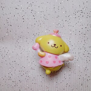 May include: A small yellow and pink plastic figurine of a dog wearing a pink dress with white wings and a pink heart-shaped wand. The figurine is on a sparkly white background.