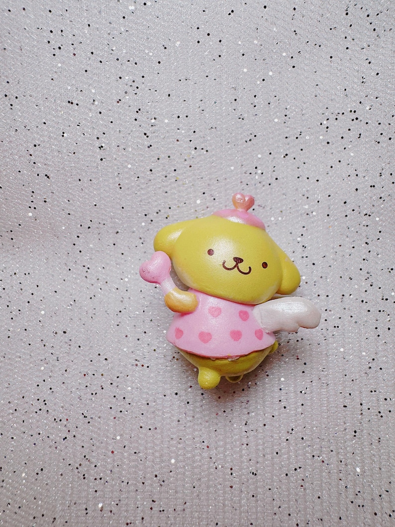 May include: A small yellow and pink plastic figurine of a dog wearing a pink dress with white wings and a pink heart-shaped wand. The figurine is on a sparkly white background.