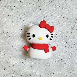 K&ouml;nnte beinhalten: Wei&szlig;e Hello Kitty Figur mit rotem Schal und Handschuhen. Die Figur hat eine rote Schleife auf dem Kopf und schwarze Augen.