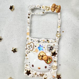 Peut inclure: Coque de t&eacute;l&eacute;phone blanche d&eacute;cor&eacute;e d'un personnage Hello Kitty, de paillettes dor&eacute;es et d'un n&oelig;ud dor&eacute;. La coque a un dos transparent et un cadre blanc.