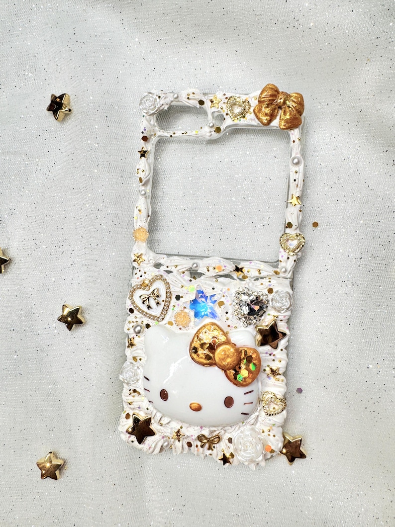 Peut inclure: Coque de t&eacute;l&eacute;phone blanche d&eacute;cor&eacute;e d'un personnage Hello Kitty, de paillettes dor&eacute;es et d'un n&oelig;ud dor&eacute;. La coque a un dos transparent et un cadre blanc.