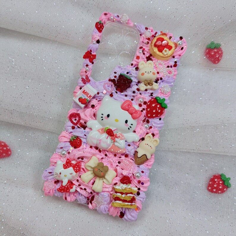 Peut inclure: Une coque de t&eacute;l&eacute;phone rose et violette d&eacute;cor&eacute;e de Hello Kitty, de fraises et d'autres d&eacute;tails mignons. La coque a un dos transparent pour montrer le t&eacute;l&eacute;phone.