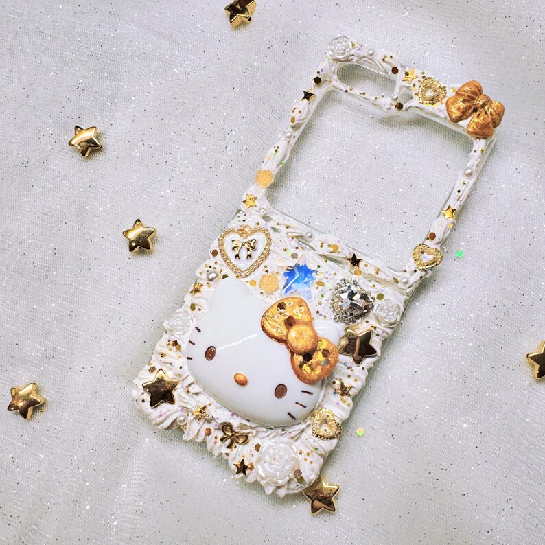 Peut inclure: &Eacute;tui de t&eacute;l&eacute;phone blanc avec un motif Hello Kitty dor&eacute;, des strass et des &eacute;toiles dor&eacute;es.
