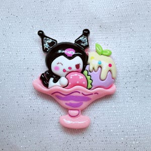 May include: A pink and purple ice cream sundae charm featuring a black and white cartoon character with a pink strawberry and a purple and yellow ice cream scoop.