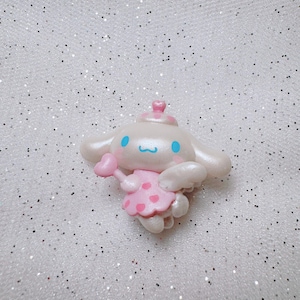 May include: A white and pink character with blue eyes and a pink heart in its hand. The character is wearing a pink dress with white polka dots and a white crown. The character is on a white sparkly background.