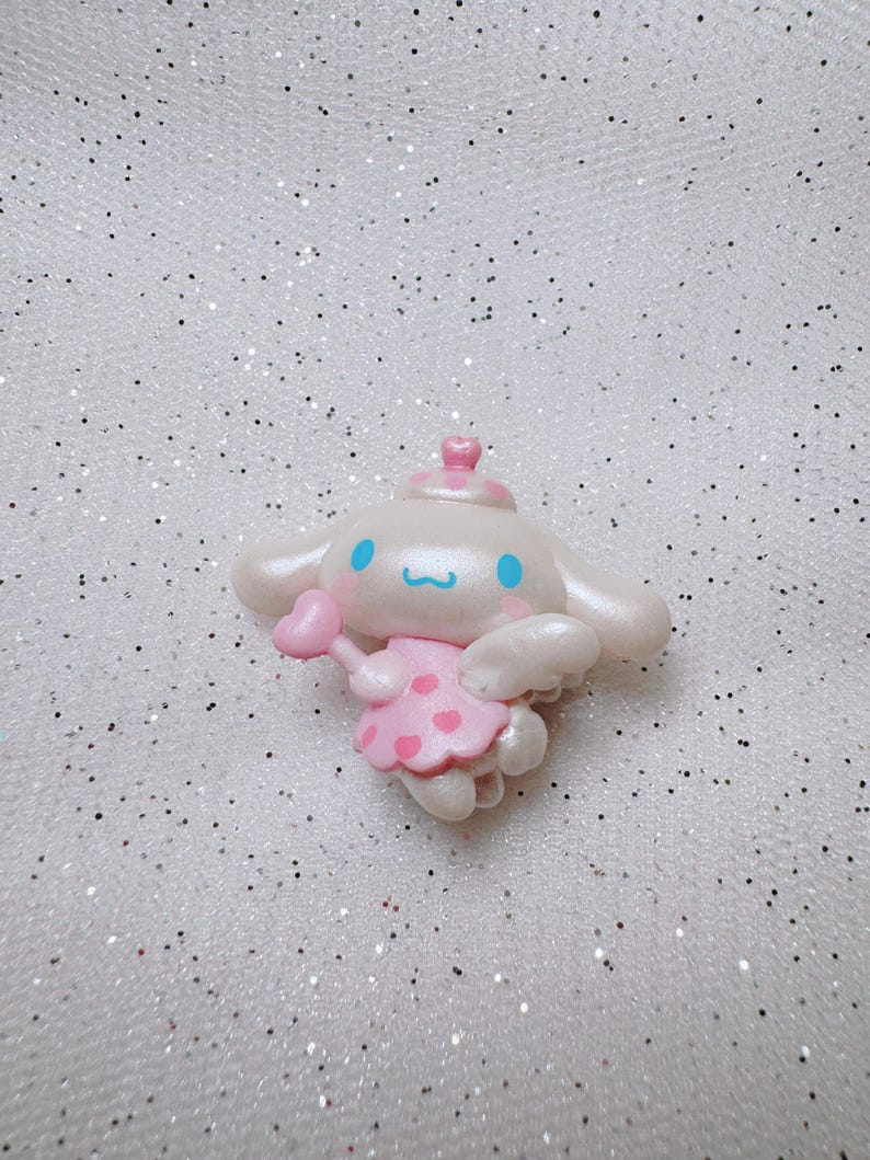 May include: A white and pink character with blue eyes and a pink heart in its hand. The character is wearing a pink dress with white polka dots and a white crown. The character is on a white sparkly background.
