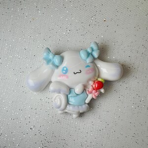 Peut inclure: Une figurine en c&eacute;ramique blanche et bleue d'un chien de dessin anim&eacute; avec un n&oelig;ud bleu, tenant une sucette rose et rouge. Le chien a un nez rose et des yeux bleus.