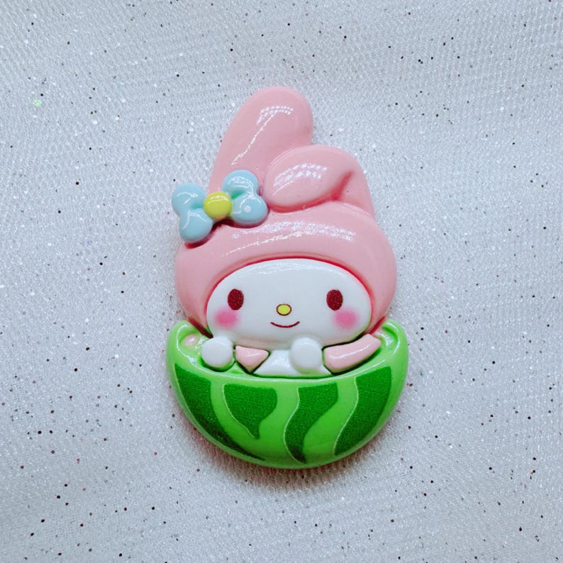 May include: A pink and white cartoon character with bunny ears and a blue bow is sitting inside a green watermelon. The character has a smiling face with closed eyes and rosy cheeks.