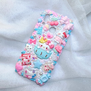 Pode incluir: Uma capa de telefone branca decorada com enfeites 3D coloridos, incluindo um personagem Hello Kitty, doces e estrelas. A capa tem um acabamento brilhante e um esquema de cores rosa e azul.