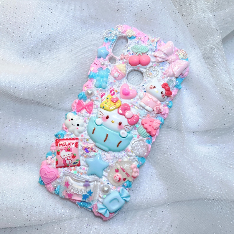 Pode incluir: Uma capa de telefone branca decorada com enfeites 3D coloridos, incluindo um personagem Hello Kitty, doces e estrelas. A capa tem um acabamento brilhante e um esquema de cores rosa e azul.