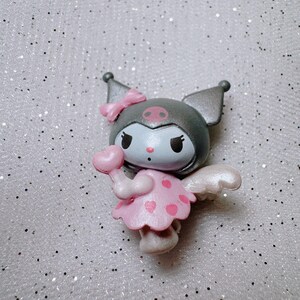 May include: A small, gray and pink figurine of a character with wings, a bow, and a heart-shaped lollipop. The character has a black face with white eyes and a pink nose.