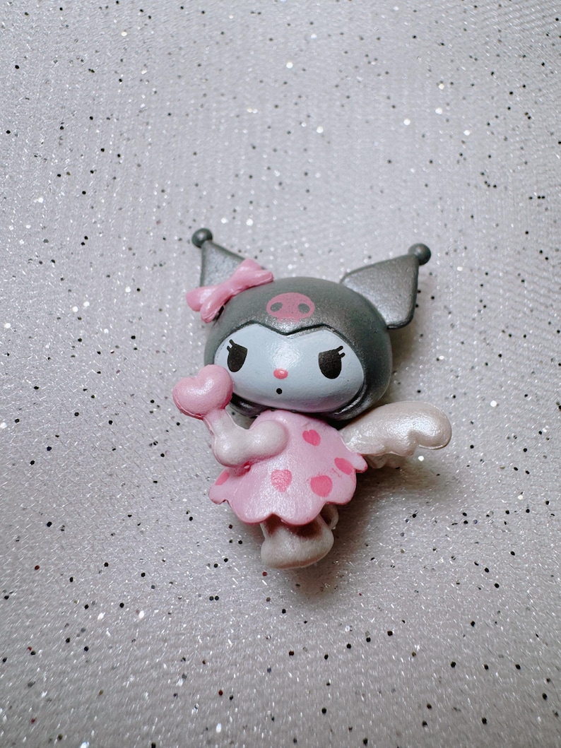 May include: A small, gray and pink figurine of a character with wings, a bow, and a heart-shaped lollipop. The character has a black face with white eyes and a pink nose.