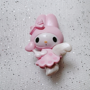 May include: Pink My Melody figurine with white wings, a pink heart-shaped wand, and a pink bow on her head. She is wearing a pink dress with white polka dots.