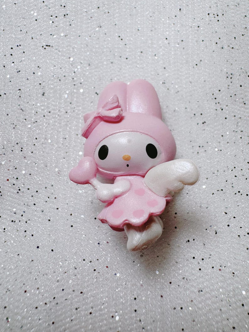 May include: Pink My Melody figurine with white wings, a pink heart-shaped wand, and a pink bow on her head. She is wearing a pink dress with white polka dots.