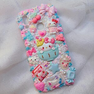 Pode incluir: Uma capa de telefone branca decorada com personagens Hello Kitty, la&ccedil;os, doces e outros detalhes fofos. A capa &eacute; coberta de glitter e tem um esquema de cores azul e rosa.
