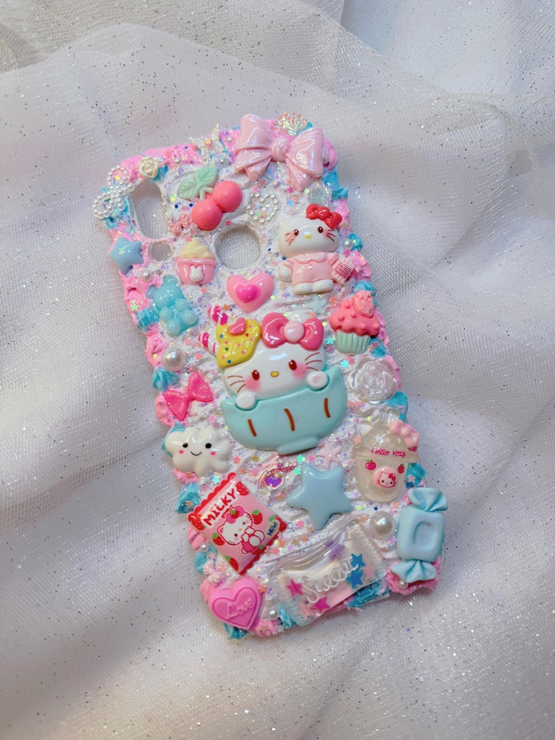 Pode incluir: Uma capa de telefone branca decorada com personagens Hello Kitty, la&ccedil;os, doces e outros detalhes fofos. A capa &eacute; coberta de glitter e tem um esquema de cores azul e rosa.