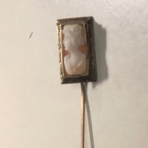 May include: A gold-tone stick pin featuring a rectangular head with a carved shell cameo. The cameo displays a white profile of a person against a peach-colored background. The metal frame surrounding the cameo has a textured, decorative border.