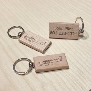 Op de afbeelding: Drie houten sleutelhangers met zilveren sleutelringen. Twee sleutelhangers hebben een gegraveerd vliegtuigontwerp, terwijl de derde de naam "John Pilot" en een telefoonnummer heeft. De sleutelhangers zijn gemaakt van lichtgekleurd hout.