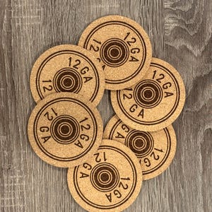 May include: A set of six round cork coasters. Each coaster has a brown, circular design with the text "12 GA" and the number "12" printed on it. The coasters are arranged on a wooden surface.