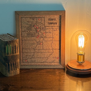 May include: A framed corkboard map of Utah with red pin markers, labeled "Utah's Temples." The map is in a wooden frame, next to a decorative wooden book and a lit Edison bulb lamp.
