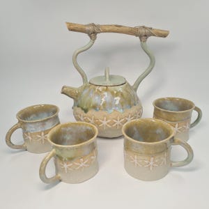 Könnte beinhalten: Ein Keramik-Teeservice mit einer Teekanne mit Holzgriff und vier passenden Tassen. Die Teekanne und die Tassen sind beige mit blauen und grünen Akzenten und einem floralen Muster. Die Teekanne hat einen Deckel und einen Ausguss.
